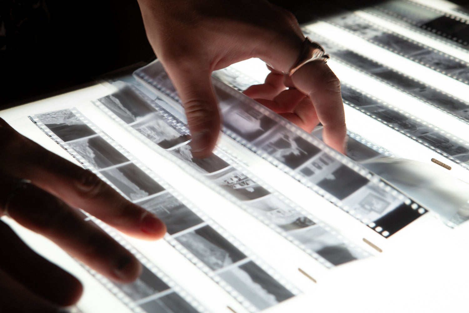 A close-up of hands examining black-and-white film strips laid out on a glowing light box in a dark room.