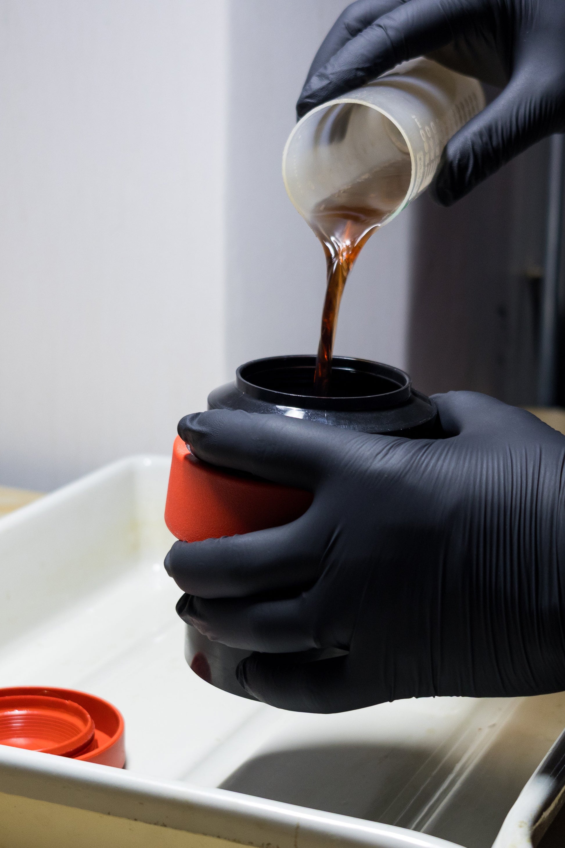 A person wearing black gloves pours a dark brown chemical from a small white cup into a black film developing tank, set over a white tray on a light wood surface.